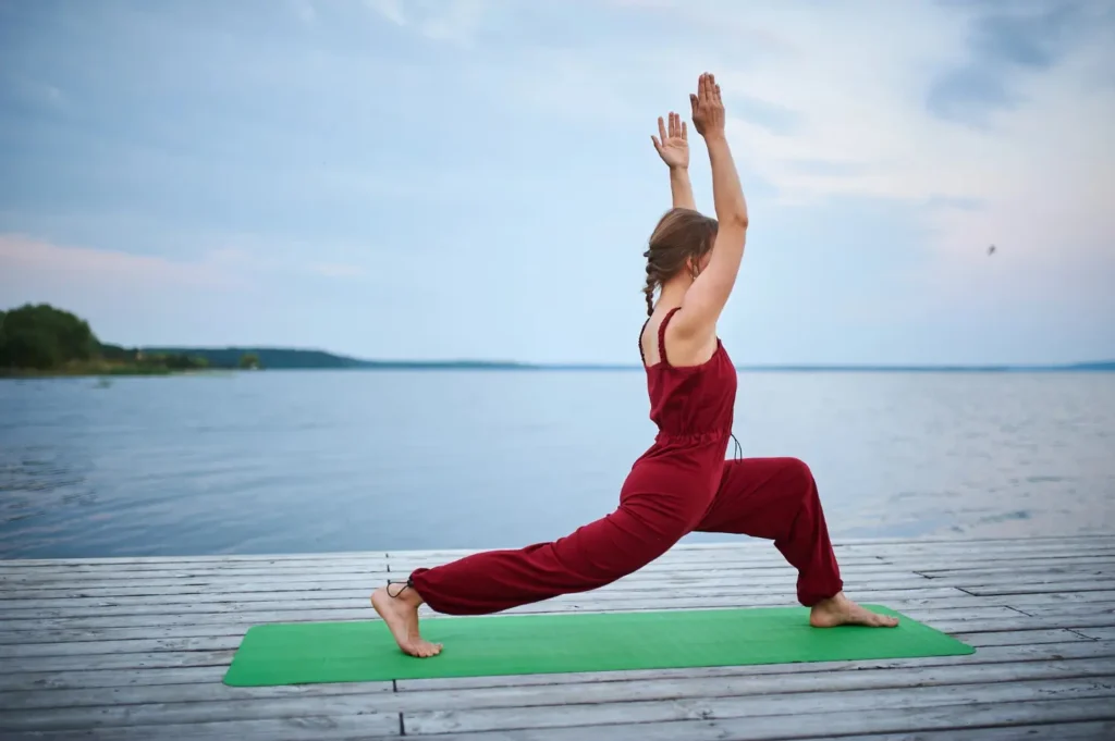 Woman practicing a yoga asana in a lunge pose on a wooden deck beside a calm lake.