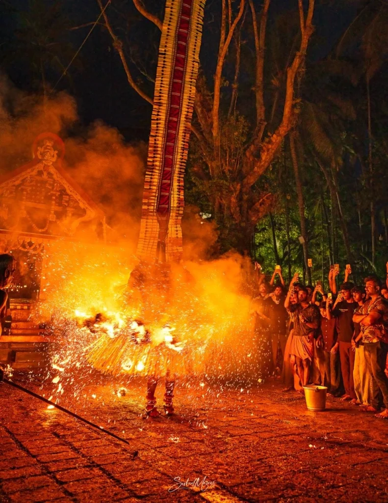 A Kandakarnan Theyyam performer wearing a towering ceremonial headdress performs a fiery ritual at night, with flames, smoke, and flying sparks illuminating the shrine courtyard while devotees watch from nearby.