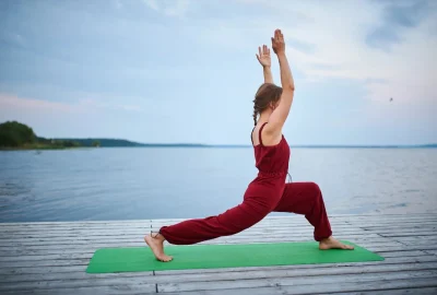 Woman practicing a yoga asana in a lunge pose on a wooden deck beside a calm lake.